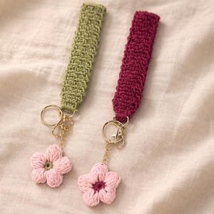 May include: Two handmade keychains with crochet flower charms. One is olive green, the other is burgundy. Both have gold-coloured hardware and pink flower charms. The keychains are displayed on a beige fabric background.