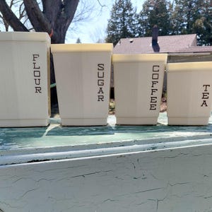 May include: Four cream-coloured kitchen canisters with lids, labelled "FLOUR", "SUGAR", "COFFEE", and "TEA" in black block letters. The canisters are rectangular with a tapered design and sit on a weathered, light blue surface.