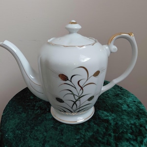 Peut inclure: Théière en porcelaine blanche avec une bordure dorée et un motif d'épis de blé. La théière a un bec verseur incurvé et une anse avec des accents dorés. Le couvercle est surmonté d'un petit bouton doré. La théière est sur une surface verte.