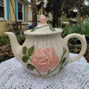 May include: Cream-colored ceramic teapot with a raised pink rose design and green leaves. The lid features a pink rosebud finial. The teapot sits on a white crocheted doily, perfect for serving tea.