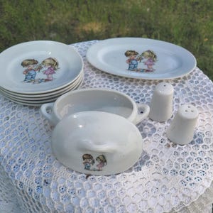 May include: A vintage white ceramic dish set with a boy and girl graphic. The set includes small plates, an oval platter, a lidded serving dish, and salt and pepper shakers. The plates measure approximately 15 cm in diameter.