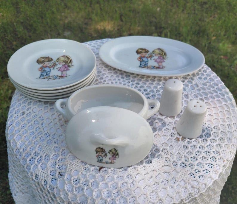 May include: A vintage white ceramic dish set with a boy and girl graphic. The set includes small plates, an oval platter, a lidded serving dish, and salt and pepper shakers. The plates measure approximately 15 cm in diameter.