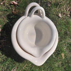 May include: Cream-colored ceramic bowl set with a speckled finish. The top bowl has two arched handles and a pouring spout. The set is nested in a larger, similarly shaped bowl. The bowls are sitting on a bed of green grass.
