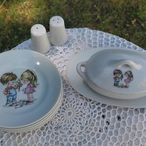 May include: A vintage white ceramic dish set featuring a child illustration. The set includes plates, a lidded serving dish, and salt and pepper shakers. The children are depicted in colourful clothing, holding flowers.