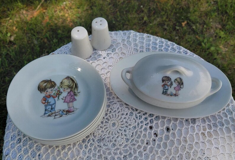 May include: A vintage white ceramic dish set featuring a child illustration. The set includes plates, a lidded serving dish, and salt and pepper shakers. The children are depicted in colourful clothing, holding flowers.