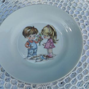 May include: A white ceramic plate with a gold rim. The plate's illustration shows a boy in blue dungarees giving flowers to a girl in a pink dress. The plate is set against a white lace background.