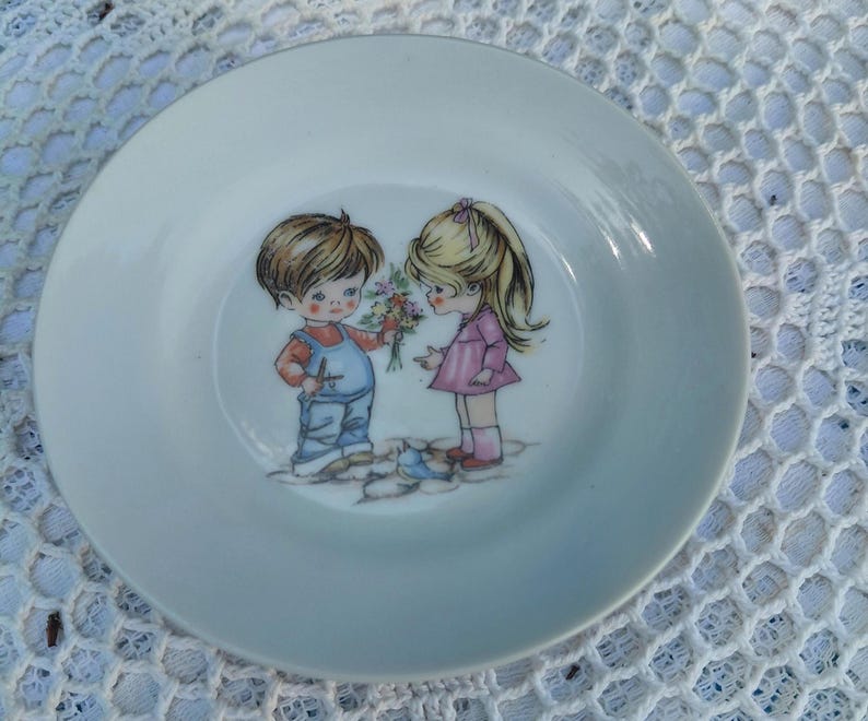 May include: A white ceramic plate with a gold rim. The plate's illustration shows a boy in blue dungarees giving flowers to a girl in a pink dress. The plate is set against a white lace background.