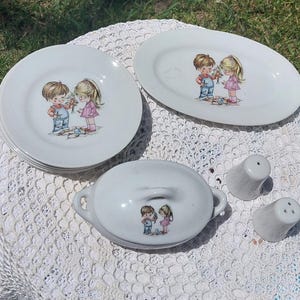 May include: A vintage white porcelain dish set with a boy and girl illustration. The set includes plates, an oval platter, a covered dish, and salt and pepper shakers. The illustration depicts a boy and girl with flowers. The set is on a white lace tablecloth.