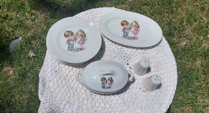 May include: A vintage white porcelain dish set with a boy and girl illustration. The set includes plates, an oval platter, a covered dish, and salt and pepper shakers. The illustration depicts a boy and girl with flowers. The set is on a white lace tablecloth.