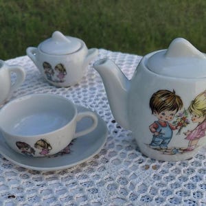 May include: A vintage white porcelain tea set with a charming illustration of a boy and girl. The set includes a teapot, creamer, sugar bowl, cup, and saucer. The teapot is approximately 15 cm tall.