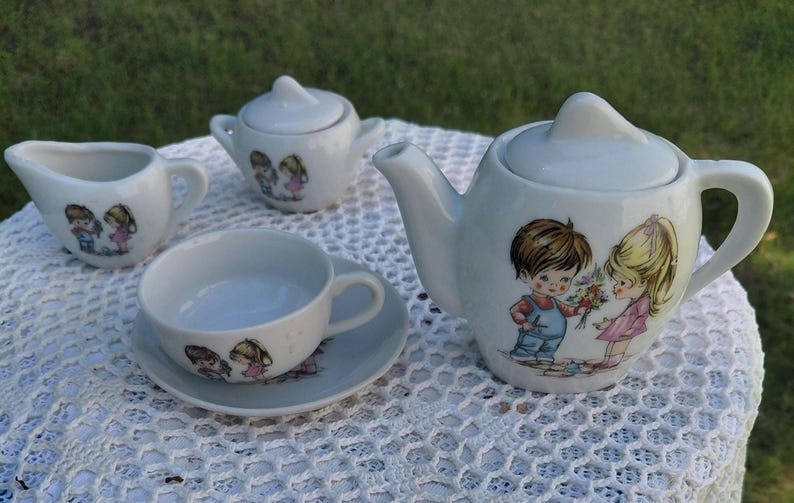 May include: A vintage white porcelain tea set with a charming illustration of a boy and girl. The set includes a teapot, creamer, sugar bowl, cup, and saucer. The teapot is approximately 15 cm tall.