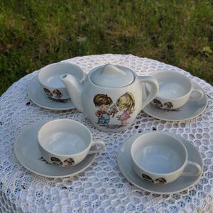 May include: A white ceramic tea set comprising a teapot, four teacups, and saucers. The teapot and saucers are decorated with a cartoon illustration of a boy and girl. The teacups and saucers have a matching design. The set is on a white lace tablecloth.