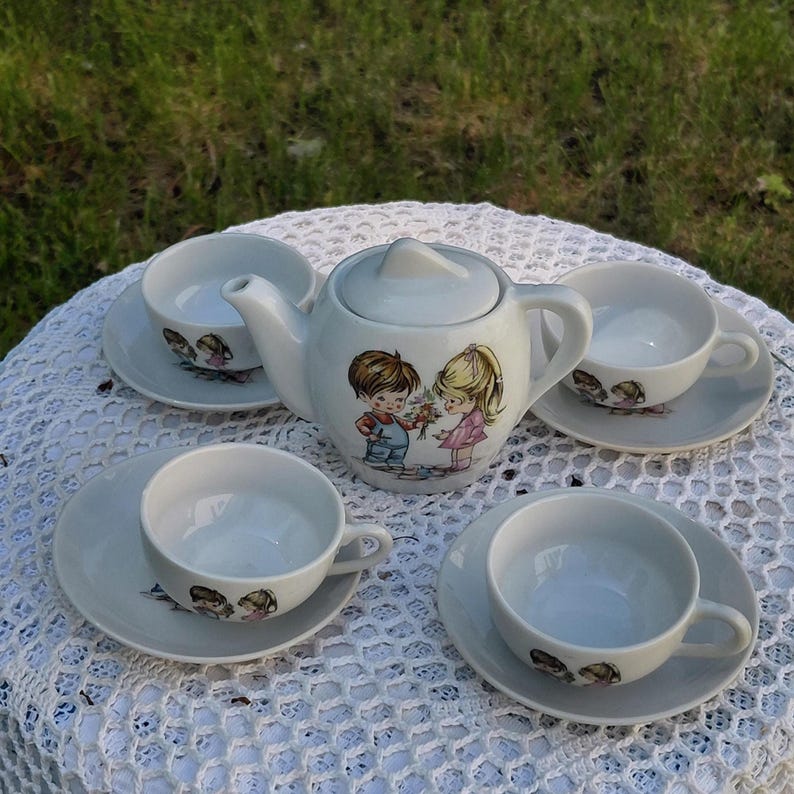 May include: A white ceramic tea set comprising a teapot, four teacups, and saucers. The teapot and saucers are decorated with a cartoon illustration of a boy and girl. The teacups and saucers have a matching design. The set is on a white lace tablecloth.