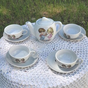 May include: A vintage white porcelain tea set comprising a teapot, four teacups, and saucers. The teapot and cups are decorated with a children's illustration. The set is arranged on a white lace tablecloth, ideal for a tea party.