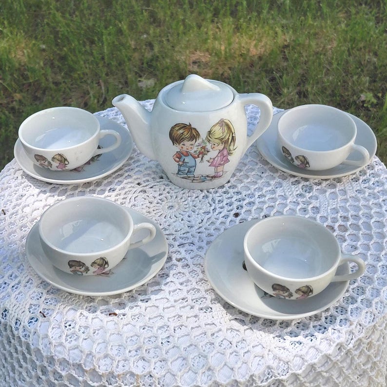 May include: A vintage white porcelain tea set comprising a teapot, four teacups, and saucers. The teapot and cups are decorated with a children's illustration. The set is arranged on a white lace tablecloth, ideal for a tea party.