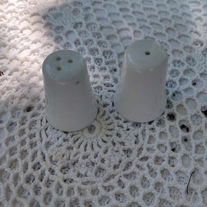 May include: Two white ceramic salt and pepper shakers. The shakers are cone-shaped with a flared base. The salt shaker has three holes, and the pepper shaker has one. They sit on a white, open-weave surface.