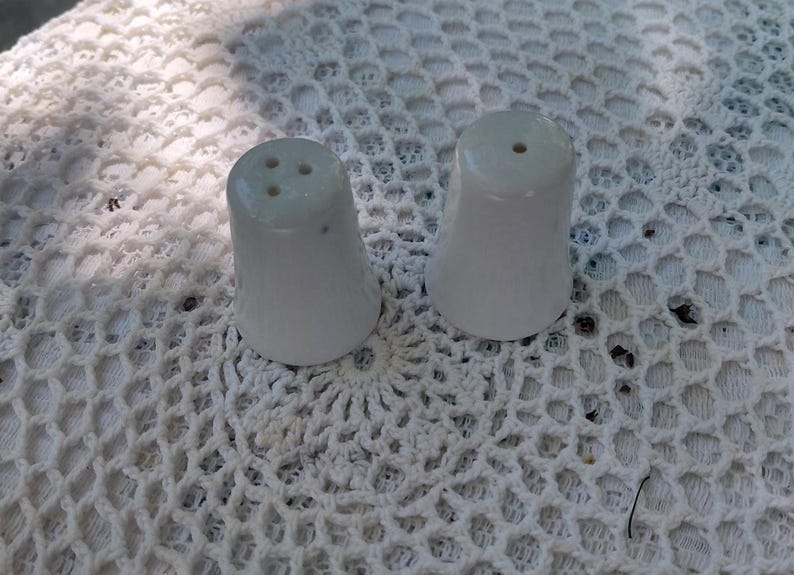 May include: Two white ceramic salt and pepper shakers. The shakers are cone-shaped with a flared base. The salt shaker has three holes, and the pepper shaker has one. They sit on a white, open-weave surface.