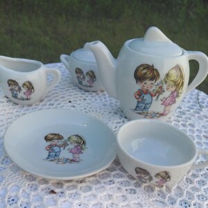 May include: A white ceramic children's tea set comprising a teapot, sugar bowl, creamer, saucer, and teacup. Each item has a cartoon illustration of a boy and girl. The teapot is the largest item.