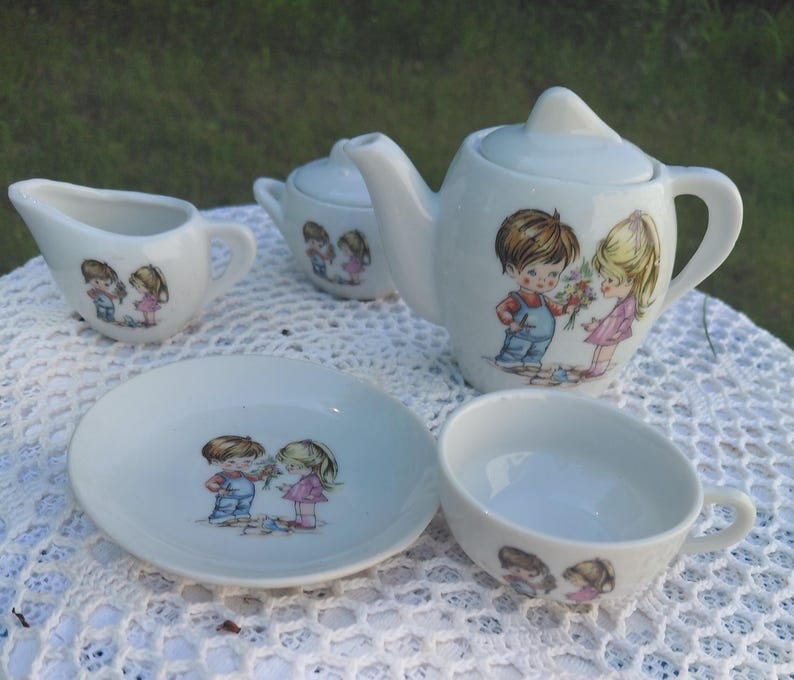 May include: A white ceramic children's tea set comprising a teapot, sugar bowl, creamer, saucer, and teacup. Each item has a cartoon illustration of a boy and girl. The teapot is the largest item.