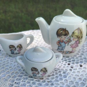 May include: A miniature white ceramic tea set comprising a teapot, creamer jug, and sugar bowl. Each item is adorned with a picture of a boy and girl. The teapot has a curved spout and handle.