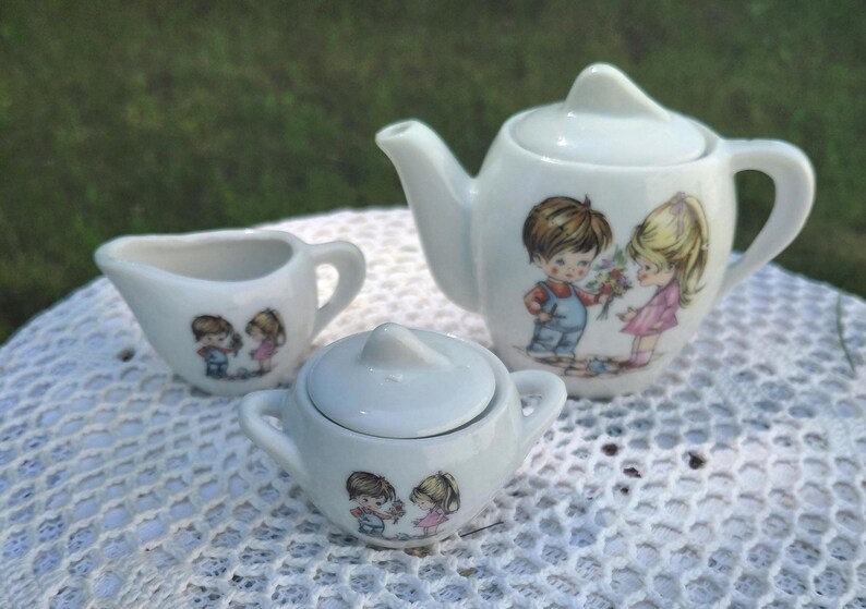 May include: A miniature white ceramic tea set comprising a teapot, creamer jug, and sugar bowl. Each item is adorned with a picture of a boy and girl. The teapot has a curved spout and handle.