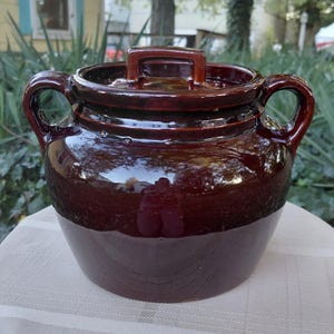 May include: A dark brown glazed ceramic bean pot with two side handles and a matching lid. The pot features a rounded body and a glossy finish, reflecting light on its surface. The lid has a central rectangular handle for easy lifting.