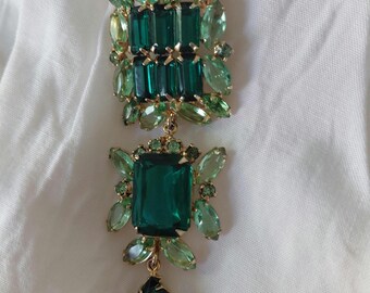 May include: A green and gold brooch. The brooch has a large rectangular emerald-cut stone at the centre, surrounded by smaller green stones. A teardrop-shaped stone hangs from the bottom, all set in a gold frame.
