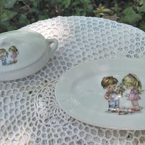 May include: A white ceramic serving dish and covered dish set. Each piece features a charming illustration of two children. The oval serving dish is approximately 25 cm long. The covered dish has two small handles.