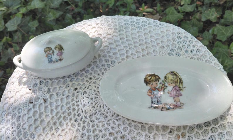 May include: A white ceramic serving dish and covered dish set. Each piece features a charming illustration of two children. The oval serving dish is approximately 25 cm long. The covered dish has two small handles.