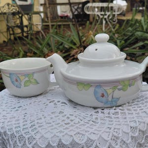 May include: White ceramic teapot and teacup set. The teapot has a lid with a small knob and a curved handle. Both pieces are decorated with a floral design in blue, green, and pink. The set is placed on a white lace tablecloth.