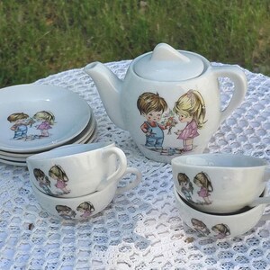 May include: A vintage white ceramic children's tea set, including a teapot, saucers, and teacups. Each item is decorated with a picture of a boy and girl. The set is displayed on a white lace tablecloth, perfect for a tea party.