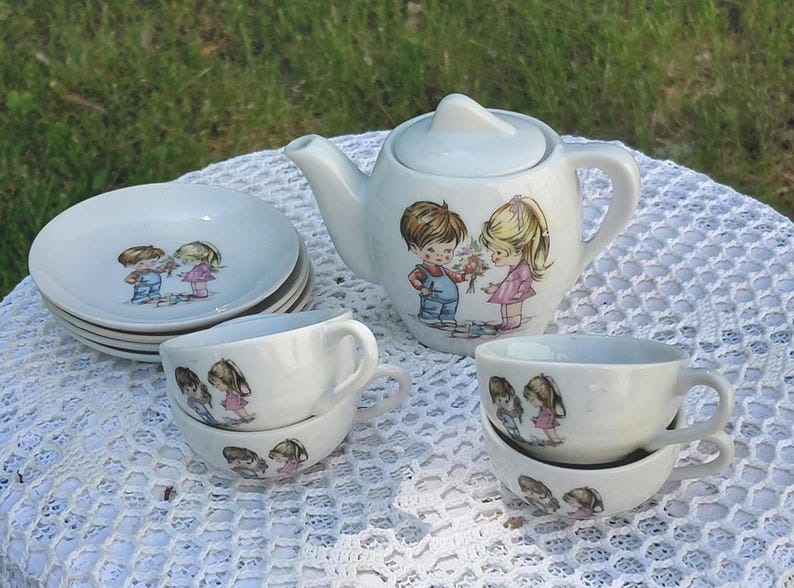 May include: A vintage white ceramic children's tea set, including a teapot, saucers, and teacups. Each item is decorated with a picture of a boy and girl. The set is displayed on a white lace tablecloth, perfect for a tea party.