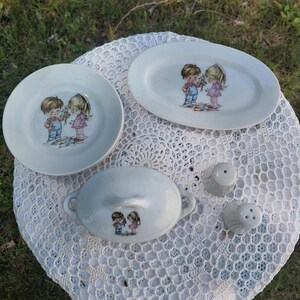May include: A collection of vintage ceramic tableware, including a round plate, an oval platter, a covered dish, and two salt shakers. Each piece features a charming illustration of a boy and girl. The items are light blue and white.