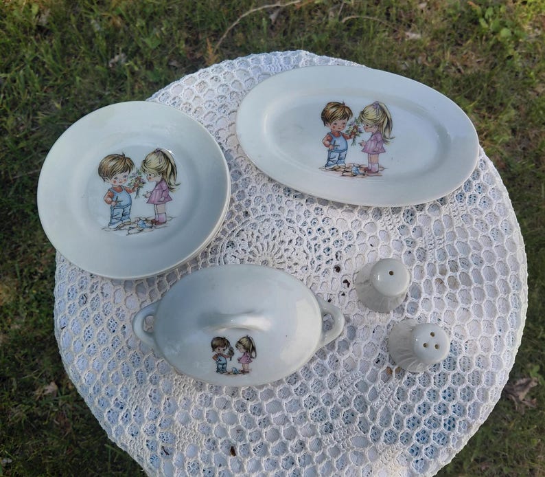 May include: A collection of vintage ceramic tableware, including a round plate, an oval platter, a covered dish, and two salt shakers. Each piece features a charming illustration of a boy and girl. The items are light blue and white.