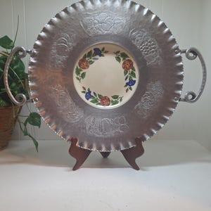 以下が含まれることがあります： 銀色の金属製サービングトレイ。エンボス加工された果物と花の模様があります。トレイはスカラップエッジと2つの湾曲したハンドルを備えています。中央には、赤、青、緑の花柄の小さな丸いプレートがあります。