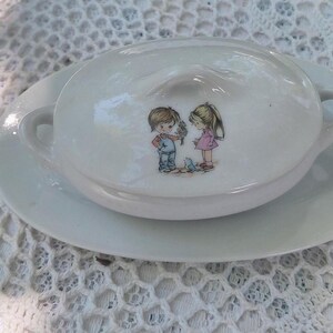 May include: White ceramic serving dish with a lid, on a matching saucer. The lid has an illustration of two children. The dish has two small handles and is on a white lace tablecloth.