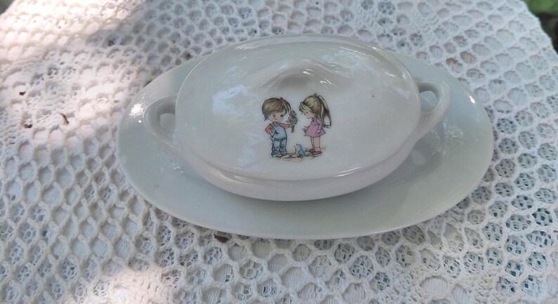 May include: White ceramic serving dish with a lid, on a matching saucer. The lid has an illustration of two children. The dish has two small handles and is on a white lace tablecloth.