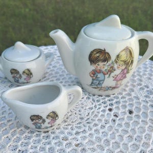 May include: A white ceramic children's tea set comprising a teapot, sugar bowl, and creamer. Each piece is decorated with a charming illustration of a boy and girl. The teapot has a lid and a handle. The set is placed on a white lace tablecloth.