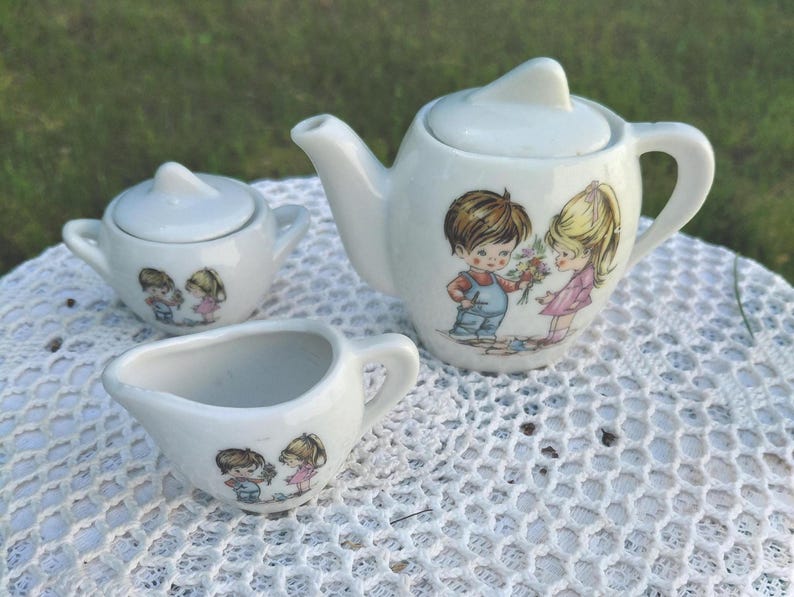 May include: A white ceramic children's tea set comprising a teapot, sugar bowl, and creamer. Each piece is decorated with a charming illustration of a boy and girl. The teapot has a lid and a handle. The set is placed on a white lace tablecloth.