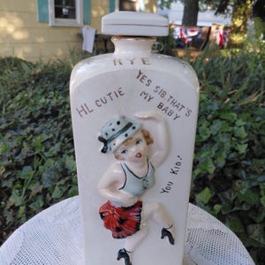 May include: A cream-colored ceramic decanter with a square stopper. The front features a raised relief of a dancing figure in a hat, blue top, and red skirt. Text on the bottle reads: RYE, HI CUTIE, YES SIR, THAT'S MY BABY, and YOU KID.