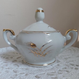 May include: White porcelain sugar bowl with a lid and two handles, accented with gold trim. The bowl features a gold wheat stalk design. The lid has a gold band and a small knob. The sugar bowl is sitting on a white lace tablecloth.