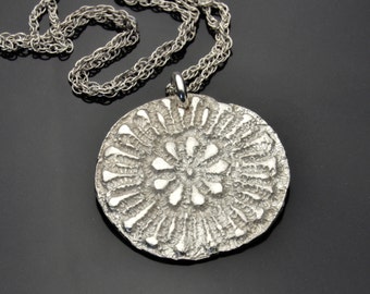 May include: A silver necklace with a round pendant featuring a textured, floral design.