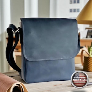 Könnte beinhalten: Eine blaue Messenger-Tasche aus Leder mit Klappenverschluss und verstellbarem Schulterriemen. Die Tasche hat eine rechteckige Form und steht auf einer Holzoberfläche. Das Bild enthält eine kleine Grafik mit der Aufschrift "Keine Zollgebühren in den USA".