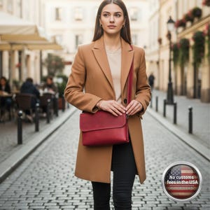 May include: A woman wearing a tan coat and holding a red leather crossbody bag. The bag has a rectangular shape with a flap closure. The woman is standing on a cobblestone street.