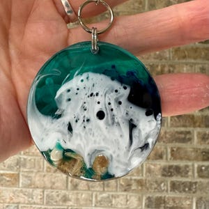 May include: A round keychain with a silver keyring and chain. The pendant features a teal, white, and black resin design resembling ocean waves. Small, tan pebbles are visible at the bottom of the pendant.