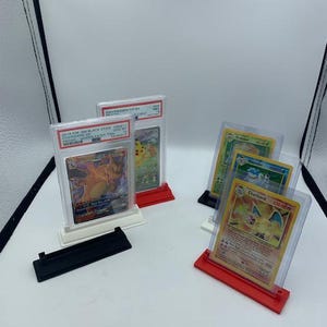 Op de afbeelding: Vier Pokémon-verzamelkaarten worden getoond in beschermende hoesjes op rode en witte acrylstandaards. De kaarten tonen Charizard, Blastoise, Venusaur en Pikachu.