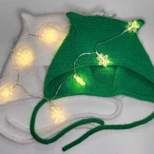 May include: Two knitted hats with ear flaps, one white and one green, decorated with a string of fairy lights. The lights are shaped like snowflakes.