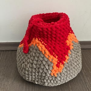 May include: A gray, orange, and red crocheted volcano-shaped storage basket. The basket has a wide opening at the top and a rounded bottom.