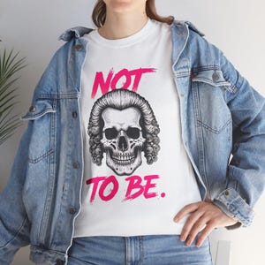Puede incluir: Camiseta blanca con un diseño de calavera con peluca empolvada y el texto rosa "NOT TO BE.". La calavera tiene detalles intrincados. La camiseta se lleva con una chaqueta vaquera azul.
