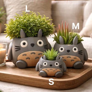 Puede incluir: Tres macetas de cerámica gris con forma de personaje de dibujos animados, cada una con una planta diferente. Las macetas están sobre una bandeja de madera, con una vela encendida a la izquierda. La maceta más grande está etiquetada como "L", la mediana como "M" y la más pequeña como "S".